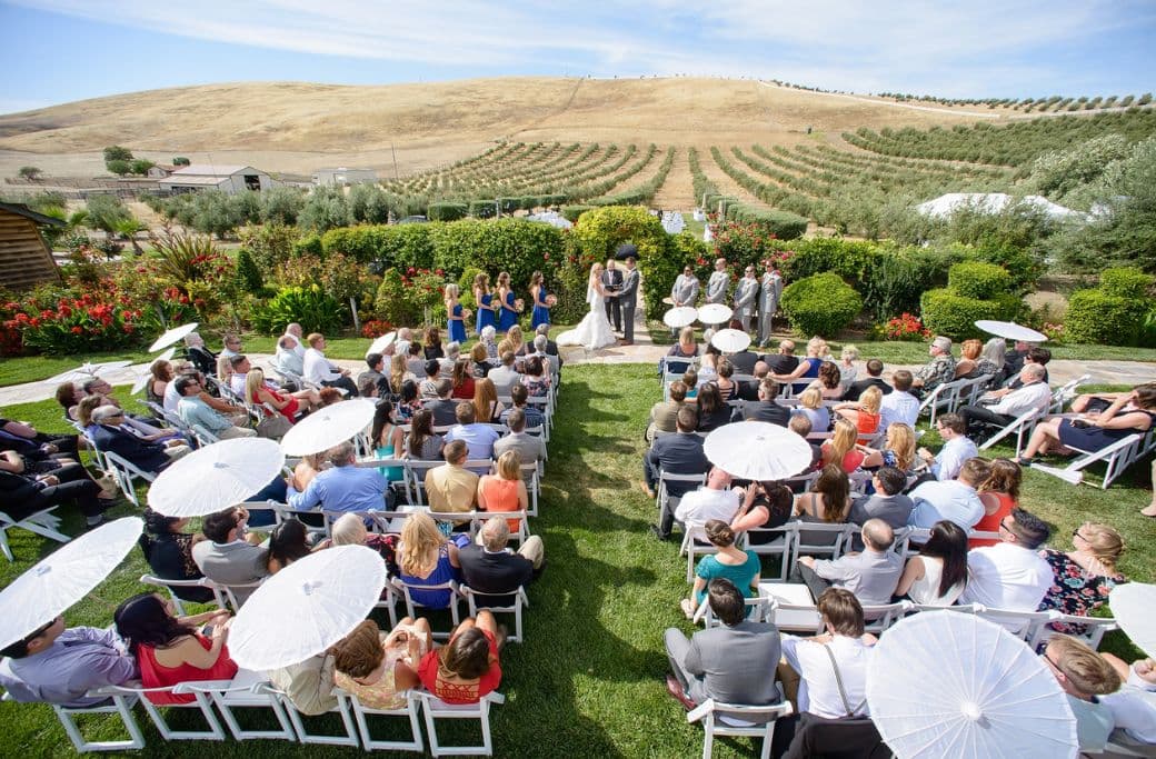 A wedding ceremony takes place outdoors in a scenic garden with guests seated among greenery and rolling hills.