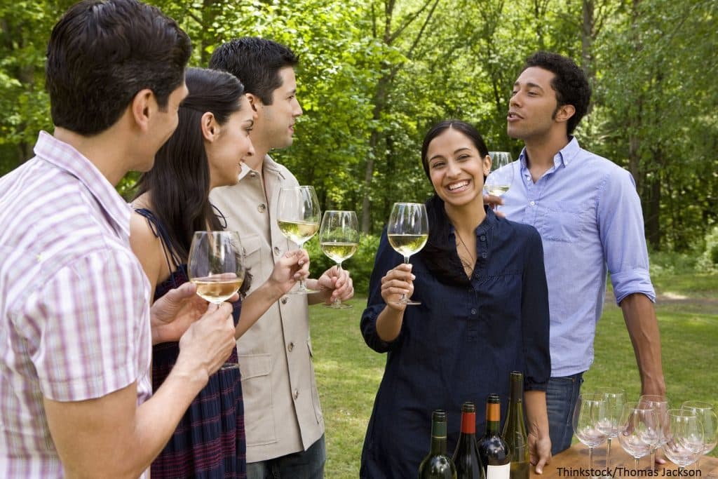 A group of five people happily toasting with glasses of white wine outdoors in a green setting. A group of five people happily toasting with glasses of white wine outdoors in a green setting.