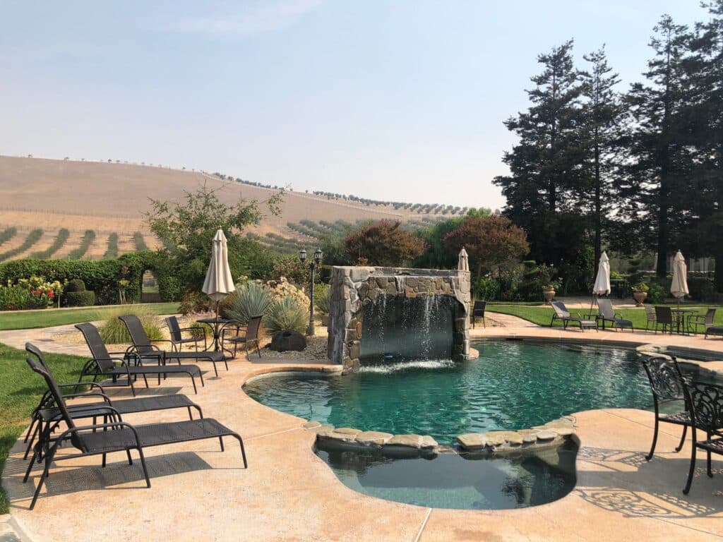 A serene pool area with a stone waterfall, surrounded by lounge chairs and lush greenery against a hillside backdrop.