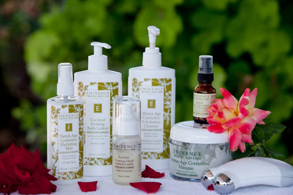 A collection of Eminence Organic Skin Care products arranged with a flower and rose petals on a blurred green background.