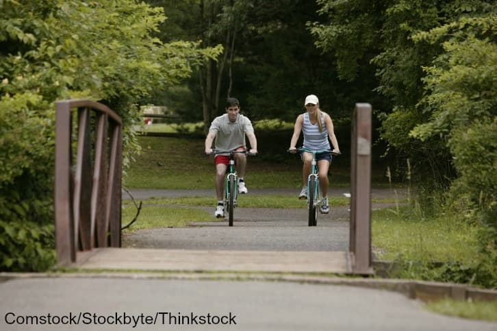 Two cyclists ride on a path near a wooden bridge surrounded by greenery.