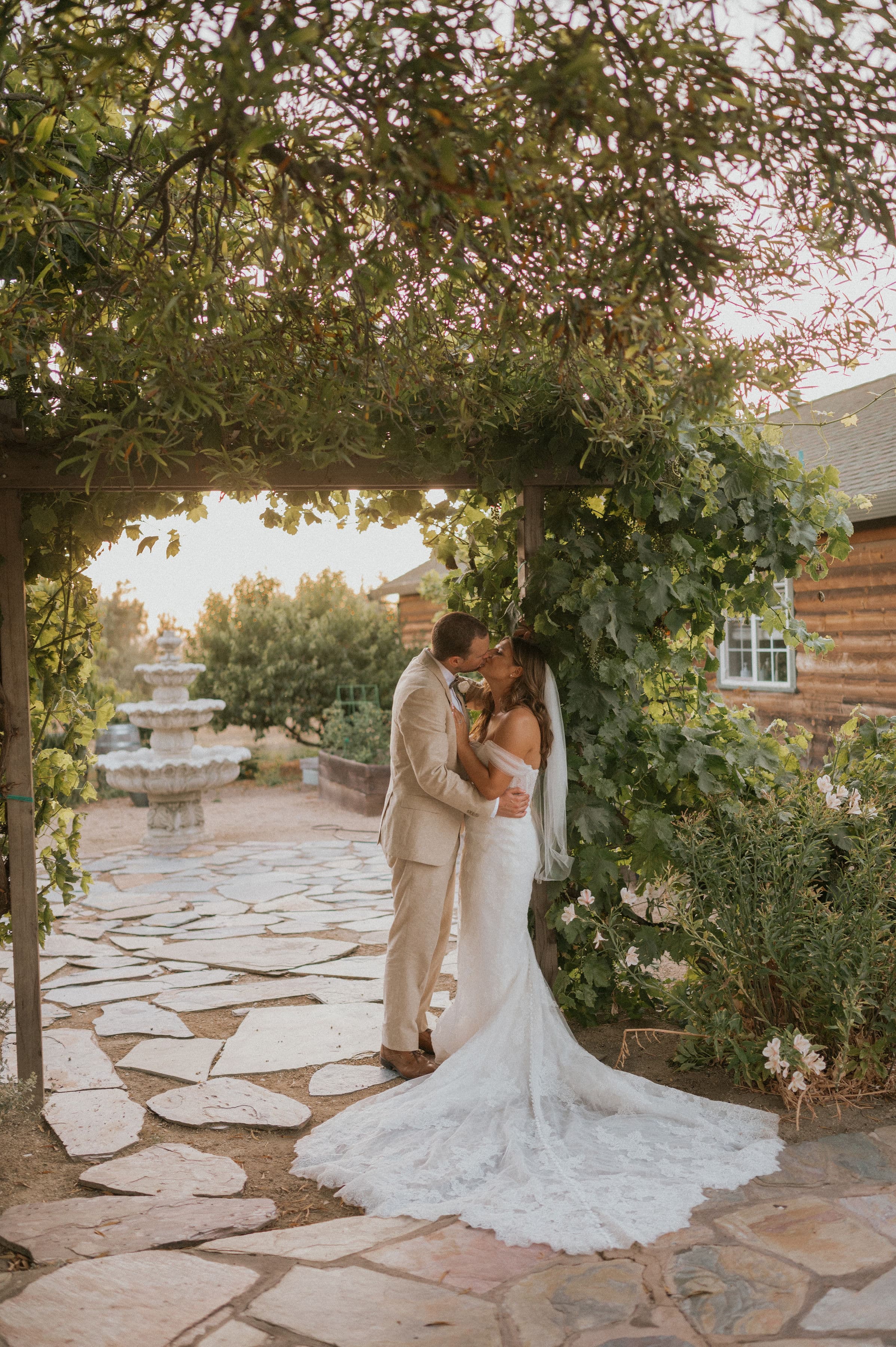 A bride and groom share a kiss under a trellis adorned with greenery in a romantic outdoor setting.