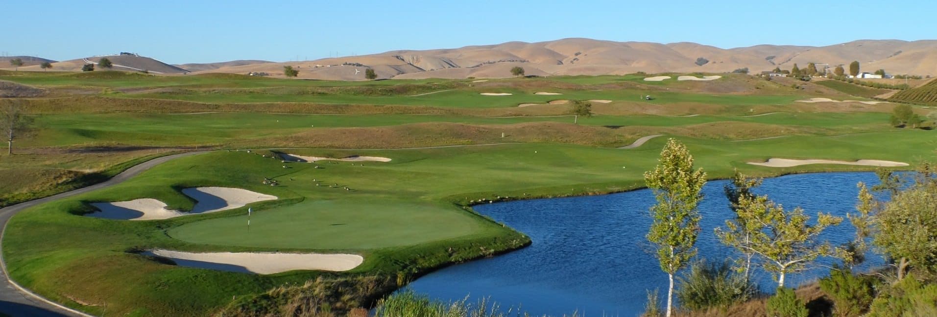 A scenic golf course featuring green fairways, sand traps, and a blue pond surrounded by rolling hills. A scenic golf course featuring green fairways, sand traps, and a blue pond surrounded by rolling hills.