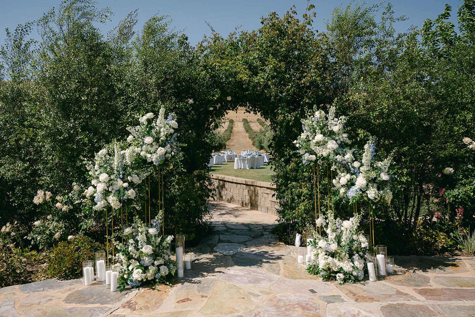 A stone pathway leads through floral arches to a reception area set with tables in a garden.