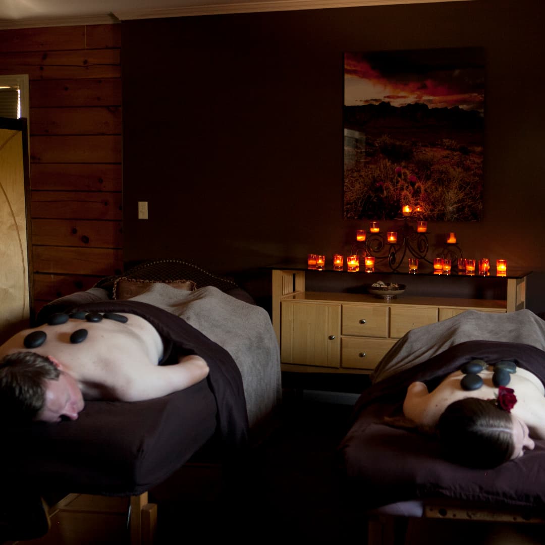 Two people receiving hot stone massages in a dimly lit spa room with candles.
