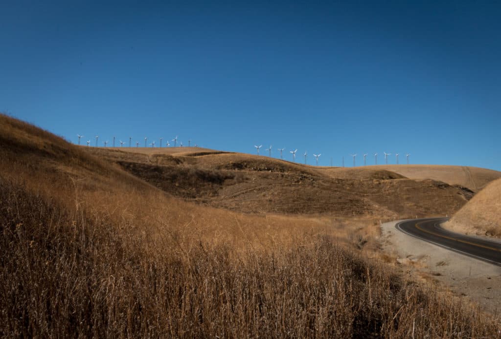 A winding road cuts through dry hills topped with wind turbines against a clear blue sky.