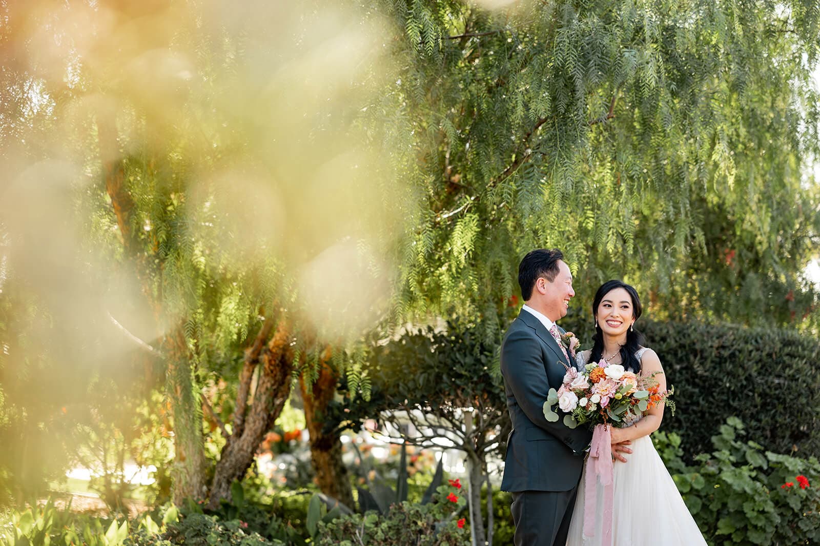 A happy couple embraces in a garden, with the bride holding a bouquet of flowers.
