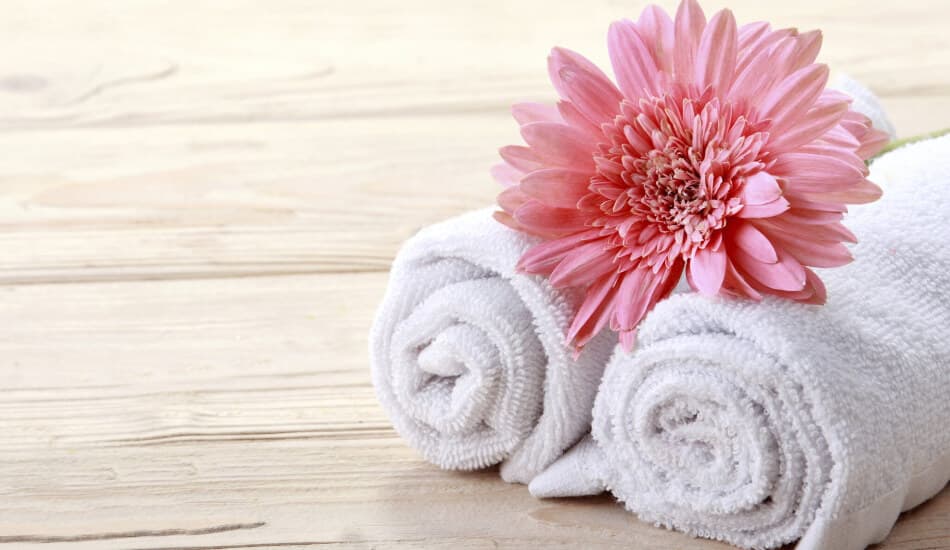 Two rolled white towels with a pink flower on top, set against a wooden background.