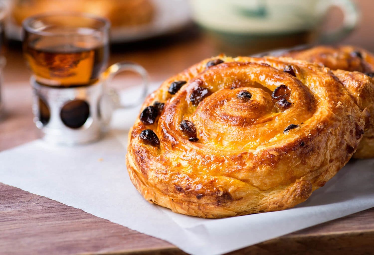 A golden-brown pastry swirled with chocolate sits on parchment paper beside a glass of tea. A golden-brown pastry swirled with chocolate sits on parchment paper beside a glass of tea.